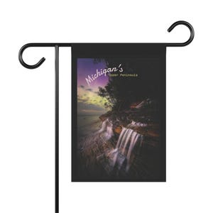 May include: Black garden flag featuring a scenic photograph of a waterfall cascading over a cliff. The flag has the text "Michigan's Upper Peninsula" in a stylized font. The flag is mounted on a black metal stand.
