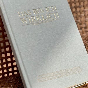 May include: A light gray journal with the text "DAS BIN ICH WIRKLICH" in gold lettering. The journal is sitting on a woven brown surface.