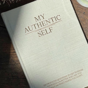 Può includere: Un diario in lino bianco con il testo "MY AUTHENTIC SELF" stampato in marrone sulla copertina anteriore. Il testo "100 PROFOUND QUESTIONS TO GET TO KNOW YOURSELF even BETTER WHO YOU REALLY are and WHAT MAKES YOU HAPPY" è stampato in marrone nella parte inferiore della copertina.