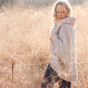 May include: A woman wearing a light beige crochet poncho with a white crocheted collar, blue jeans, and white socks. She is standing in a field of tall, dry grass.