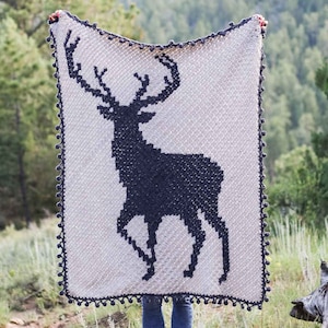 May include: A crocheted blanket with a navy blue deer silhouette on a white background. The blanket has a navy blue border with pom-poms.