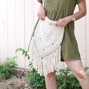 Pode incluir: Uma mulher vestindo um vestido verde, um chapéu marrom e óculos de sol está segurando uma bolsa branca de crochê com franjas. Ela está em pé na frente de uma parede de madeira branca.