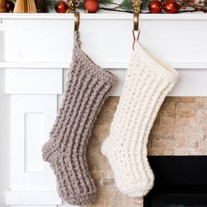 May include: Two crocheted Christmas stockings hanging on a white fireplace mantel. The stocking on the left is a light brown color and the stocking on the right is white.