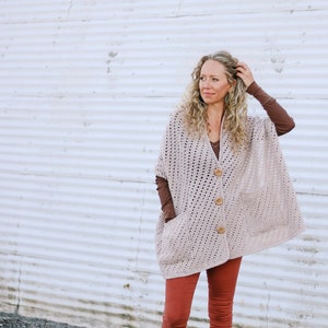 Può includere: Una donna indossa un poncho in crochet color marrone chiaro con chiusura a bottone e tasche. Indossa pantaloni rossi e stivali marroni.