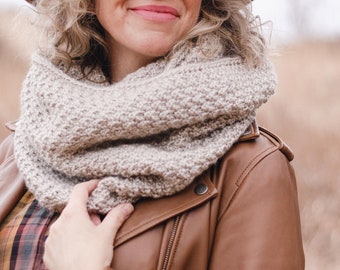 Könnte beinhalten: Eine Frau trägt eine braune Lederjacke, einen beigefarbenen Strick Schal und einen braunen Fedora Hut. Sie schaut zur Seite und lächelt.