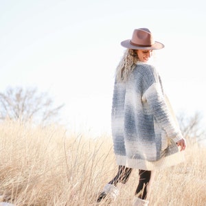 以下が含まれることがあります： 茶色のフェドーラハットをかぶった、グレーとホワイトのクロシェカーディガンを着た女性が、背の高い乾いた草のフィールドに立っています。