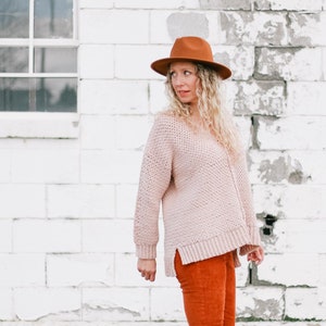 May include: A woman wearing a light pink sweater, rust-colored pants, and brown clogs. She is wearing a brown fedora hat and has curly blonde hair.