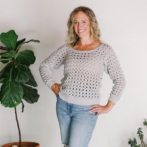 May include: A woman wearing a light grey crochet sweater with a loose fit and a square pattern. She is wearing light blue jeans with a distressed look and brown sandals with buckles.