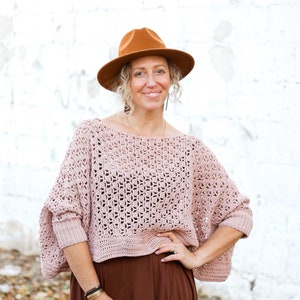 Könnte beinhalten: Eine Frau trägt einen hellrosa gehäkelten Pullover mit einem braunen Hut und einem braunen Rock. Sie steht vor einer weißen Backsteinwand.