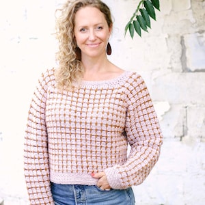 May include: A light pink and brown crochet sweater with a square pattern. The sweater has long sleeves and a cropped fit. The model is wearing blue jeans.