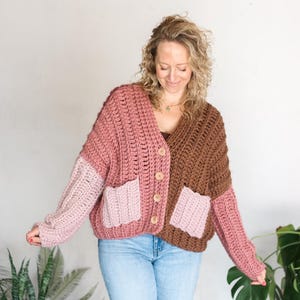 May include: A chunky, oversized crochet cardigan in shades of pink and brown. The sweater features a color-blocked design with two front pockets and wooden buttons. The sleeves are a lighter shade of pink.