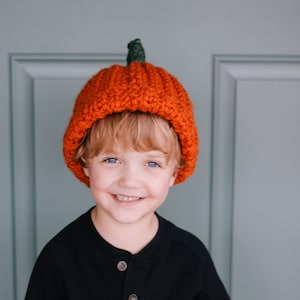 May include: A young child wearing a black long-sleeved shirt with a single button at the neck. The child is wearing a knitted orange pumpkin hat with a green stem.