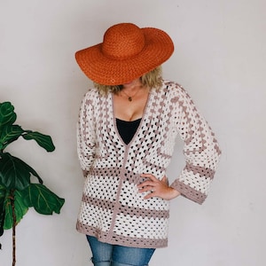 Op de afbeelding: Een vrouw draagt een gehaakt vest met witte en bruine strepen, een spijkerbroekje en een brede oranje zonnehoed.