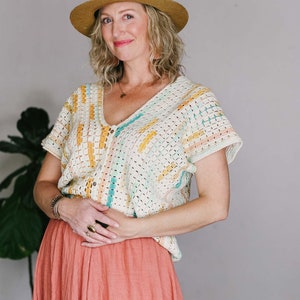 May include: A woman wearing a crocheted top with a colourful striped pattern and a wide-brimmed hat. She is wearing a coral-coloured skirt and has her hands clasped in front of her.