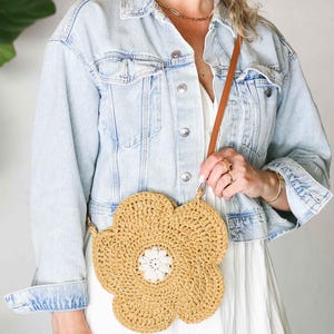 May include: A round, flower-shaped crochet crossbody bag in a tan, straw-like yarn with a white, textured center. The bag features a thin, brown leather-like strap attached to the top, creating a distinct floral silhouette against a light outfit.