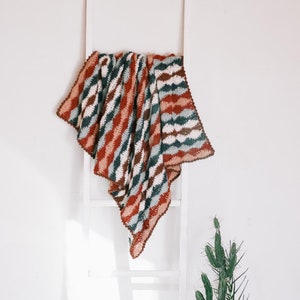 May include: A white ladder with a crocheted blanket draped over it. The blanket has a wavy pattern in shades of brown, red, and blue. A cactus plant in a woven pot sits on the floor next to the ladder.