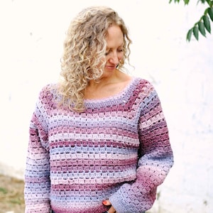 May include: A handmade, crochet sweater in shades of purple, lavender, and blue. The sweater has a square neckline and long sleeves. The model is wearing blue jeans. The sweater is a cozy, handmade garment.