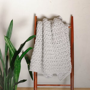 May include: A light gray crocheted throw blanket draped over a wooden ladder style stand. The stand has two rungs and is made of dark wood. A green plant is in a terracotta pot in the foreground.