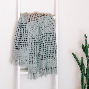 Pode incluir: Uma manta cinza claro de crochê com um padrão quadrado e franjas está pendurada em uma escada de madeira branca. Um cacto em vaso está à direita da escada contra uma parede branca.