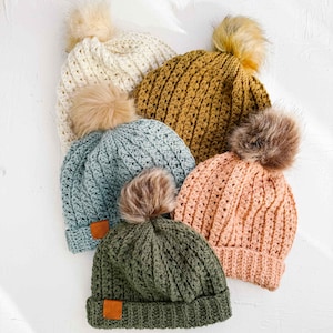 May include: A collection of six handmade crochet beanies in various colors, including cream, mustard yellow, dusty blue, peach, and olive green. Each hat features a faux fur pom-pom and a small brown leather tag.