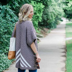 Peut inclure: Une femme portant un cardigan en crochet gris avec des accents en chevron blanc, un jean bleu et des sandales brunes. Elle porte un sac fourre-tout brun et blanc.