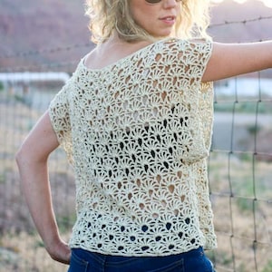 May include: A woman wearing a crocheted top with a delicate openwork pattern. The top is a light beige colour and has short sleeves. The woman is wearing blue jeans and has her hair pulled back.