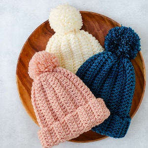 Three white, pink and blue easy adult beanies with pom poms.