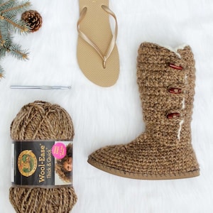 May include: A pair of brown crochet boots with brown buttons and a white fleece lining. The boots are on a white fuzzy surface. There is a ball of brown yarn, a crochet hook, a pine cone, and a pair of flip flops in the image. The yarn label reads "Wool-Ease Thick & Quick".