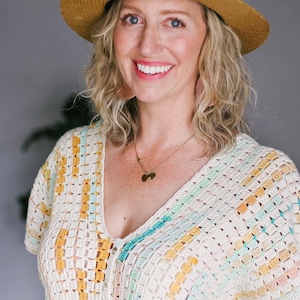 A blonde woman looking looking at the camera and smiling. She is wearing a v neck crochet rectangle top and a tan hat.