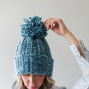 A blonde woman wearing a blue 1.5 hour beanie with her hand on the pompom.