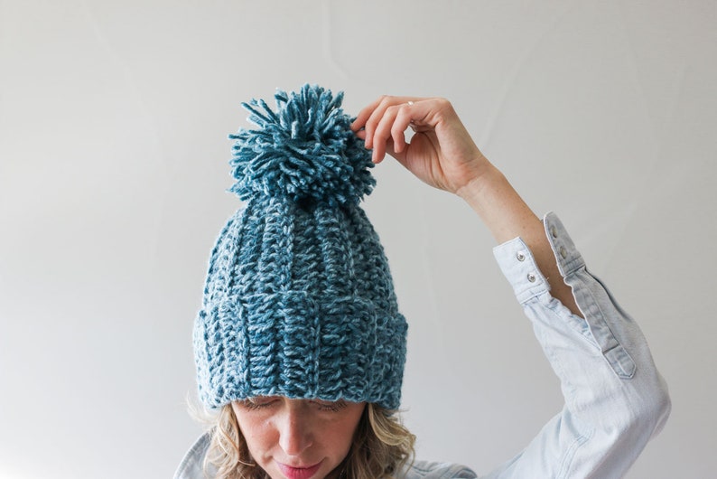 A blonde woman wearing a blue 1.5 hour beanie with her hand on the pompom.