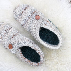 A pair of white house shoes with a button detail placed on a white fur rug.