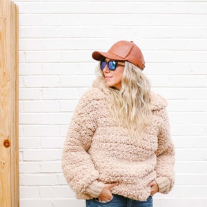 A blonde woman standing, looking sideways, wearing a brown cap, a pair of sunglasses, a beige oversized sweater with a hood, and denim jeans with her hands on the front pockets of her jeans.