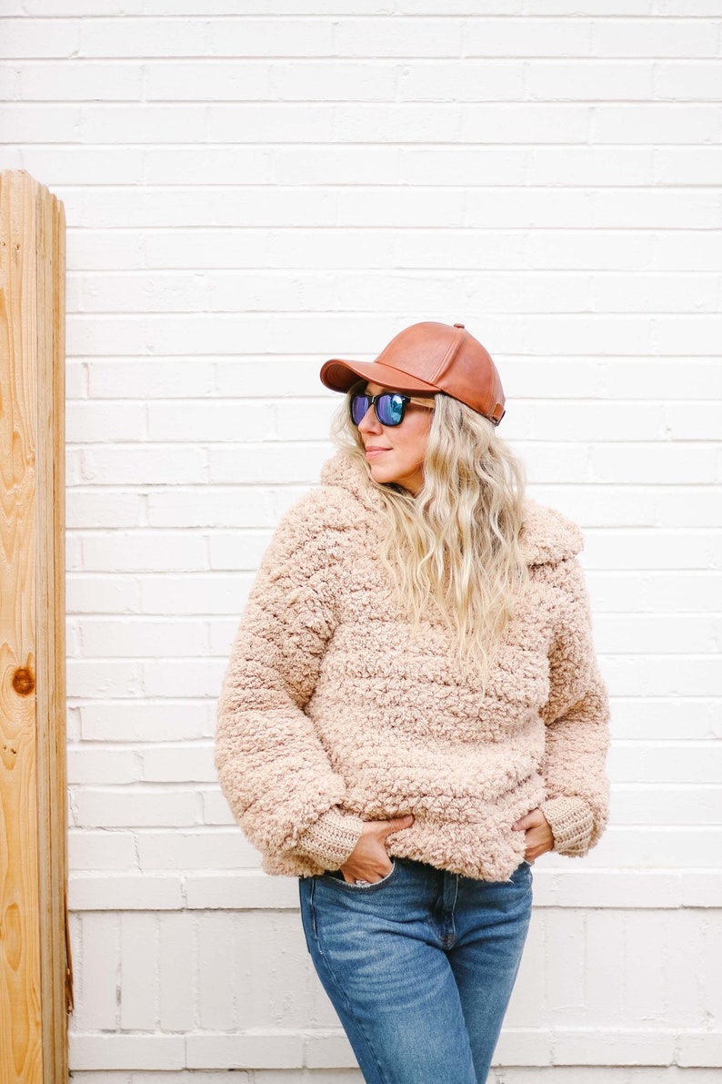 A blonde woman standing, looking sideways, wearing a brown cap, a pair of sunglasses, a beige oversized sweater with a hood, and denim jeans with her hands on the front pockets of her jeans.