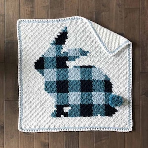May include: A white crocheted baby blanket with a blue and black plaid bunny design. The blanket has a blue border and a white pom-pom.