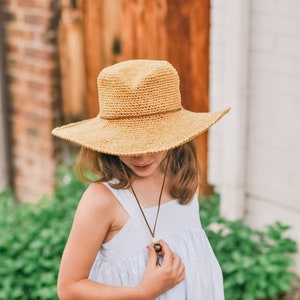 A little girl looking down, smiling wearing a brown wide brim fedora hat and a baby blue dress with her hand on the hat's adjuster.