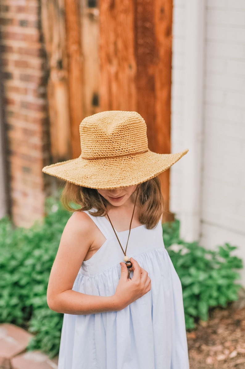 A little girl looking down, smiling wearing a brown wide brim fedora hat and a baby blue dress with her hand on the hat's adjuster.