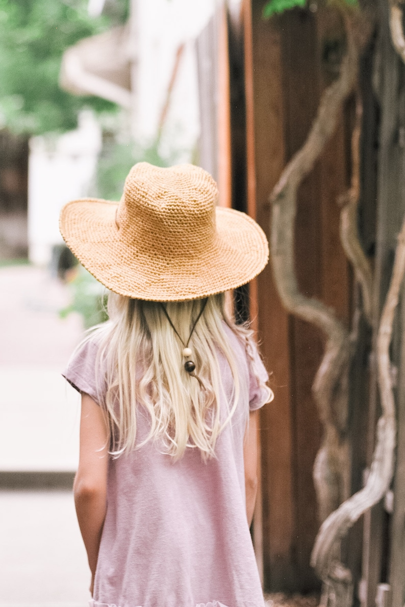 A blonde little girl with her back on the camera wearing a brown kids sun hat and pink t-shirt dress.