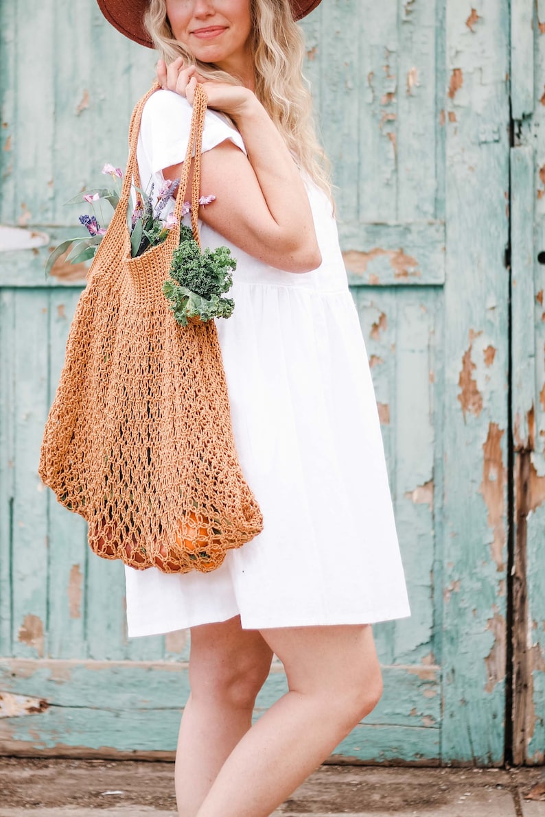 A blonde woman is carrying an easy-tote bag full of vegetables, fruits, and flowers.