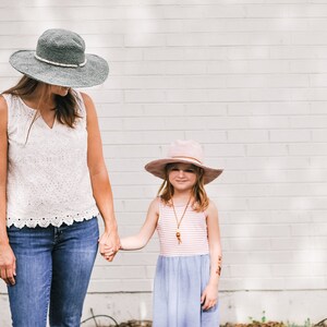 A brunette woman holds the hand of a little girl wearing a matching crochet sun hat, the woman wearing a green one, and the little girl wearing a pink one.