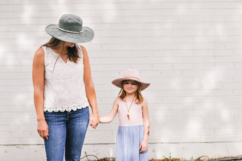 A brunette woman holds the hand of a little girl wearing a matching crochet sun hat, the woman wearing a green one, and the little girl wearing a pink one.