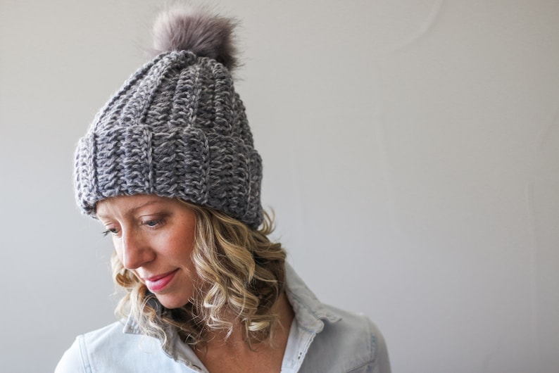 A blonde woman wearing a gray 1.5 hour crochet hat with pom poms and a denim polo top.