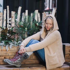 May include: A woman wearing a beige hooded cardigan and blue jeans sits on a wooden bench. She is wearing brown boots with red laces and a white fur trim. The background features a wall of birch branches and greenery.