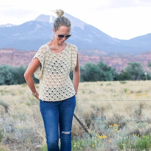 A blonde woman standing on a field, looking down, smiling, wearing a pair of sunglasses, a crochet summer top, a black bandeau, and denim jeans.
