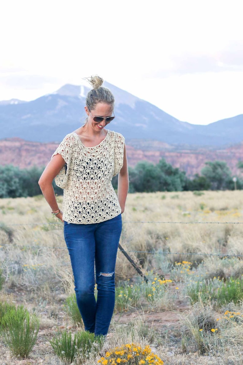 A blonde woman standing on a field, looking down, smiling, wearing a pair of sunglasses, a crochet summer top, a black bandeau, and denim jeans.