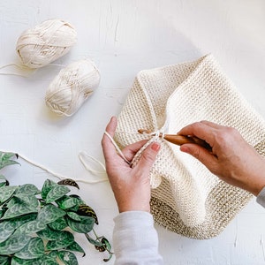 A woman crocheting an in-progress crochet backpack using a crochet hook with two yarn skeins.