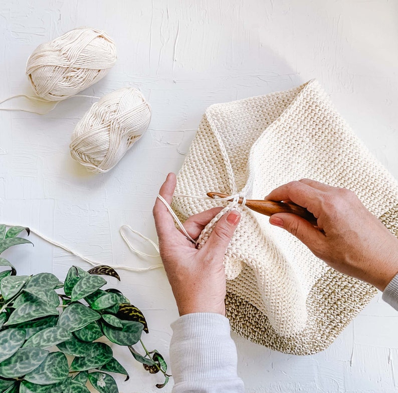 A woman crocheting an in-progress crochet backpack using a crochet hook with two yarn skeins.