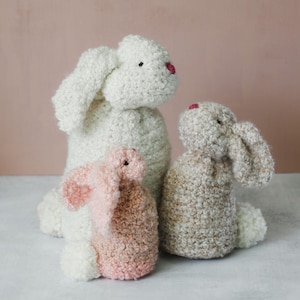 May include: Three crocheted bunny plushies in different sizes and colors. The largest bunny is white, the middle bunny is beige, and the smallest bunny is pink.