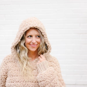 A blonde woman smiling, wearing a beige women's crochet hoodie with the hood on her head with her hand holding the hood.