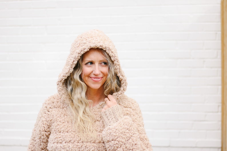 A blonde woman smiling, wearing a beige women's crochet hoodie with the hood on her head with her hand holding the hood.